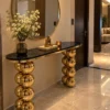 Black marble console table with stacked gold sphere legs placed beneath a round mirror in a luxury home entryway.