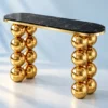 Gold stainless steel console table with black marble rounded rectangular top and stacked spherical legs against a pale blue background