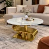 Designer round marble centre table with gold stainless steel curved base displayed in front of a modern beige sectional sofa.