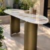 Elongated oval white marble console table with bronze-gold curved stainless steel supports in a sunlit garden courtyard.