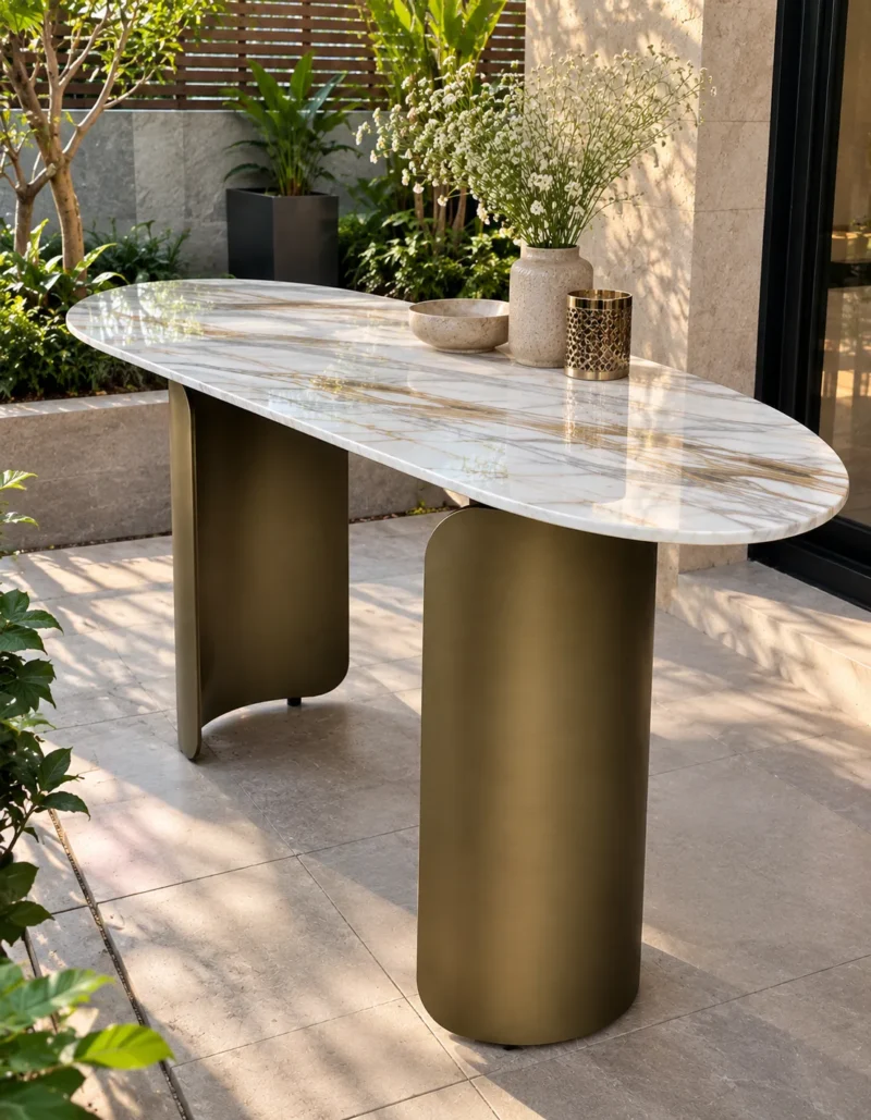 Elongated oval white marble console table with bronze-gold curved stainless steel supports in a sunlit garden courtyard.