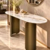 Luxury marble console table with elongated oval top and bronze-gold stainless steel base beneath a round wall mirror.