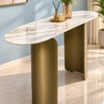 Modern oval marble console table with curved bronze stainless steel pedestal base styled with a vase and decorative tray against a pale blue wall.