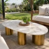 Oval white marble coffee table with three gold stainless steel pedestal legs beside a cushioned sofa on a garden patio.