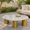 White oval marble centre table with grey and gold veining supported by three cylindrical gold stainless steel legs in a sunny patio setting.