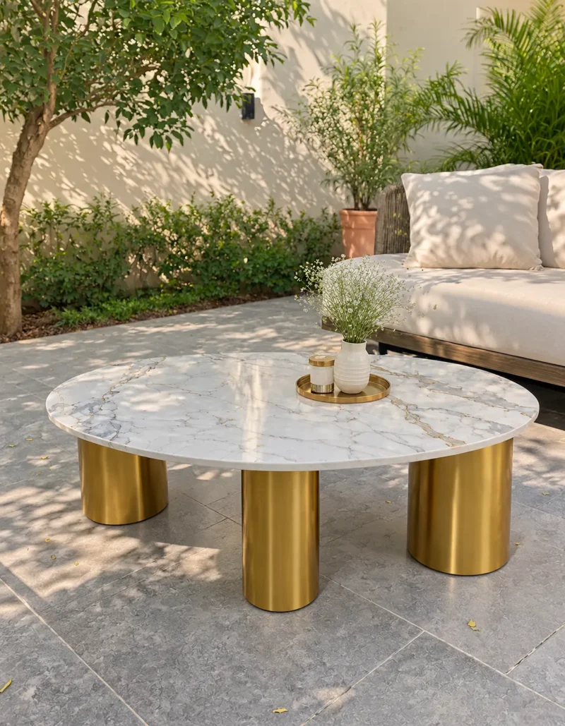 White oval marble centre table with grey and gold veining supported by three cylindrical gold stainless steel legs in a sunny patio setting.