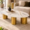 Modern oval white marble coffee table with gold stainless steel cylindrical base in front of a beige upholstered sofa.