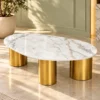 Designer oval white marble centre table with three gold cylindrical stainless steel pedestals in a sunlit minimalist interior.