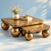 Luxury gold nesting coffee table set with rounded square tabletops and spherical legs styled against a pale blue studio background.
