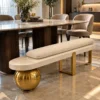 Luxury ivory bouclé upholstered bench with gold sphere and stainless steel legs positioned beside a marble dining table.