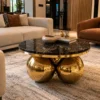 Round black marble centre table with polished gold sphere base between cream and brown sofas in a luxury living room.