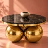 Round black marble centre table with gold sphere base photographed against a warm terracotta studio background.