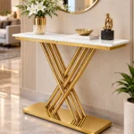 Gold stainless steel console table with white marble top, geometric crossed rod base and decorative vase beneath a circular mirror in a luxury living room.