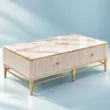 White marble centre table with gold PVD stainless steel frame, ribbed storage body and rectangular top against a light blue studio background.
