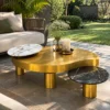 Sculptural gold coffee table with raised white marble accent and lower black marble surface beside a cream sofa in a garden lounge.