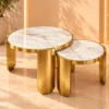 Two round white marble nesting tables with gold stainless steel frames against a warm cream studio background.