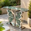 Green marble butterfly console table with polished silver stainless steel base displayed in a sunlit garden patio setting.