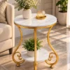 Designer gold scrollwork marble side table beside a cream sofa with flowers and a plant on its two marble tiers