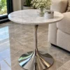 Round white marble side table with reflective silver chrome tulip pedestal beside a luxury sofa