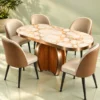 Oval marble dining table with gold-trimmed white tabletop, sculptural wood-look pedestal and cream-and-caramel upholstered chairs against a green background.