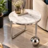 Designer marble side table with silver stainless steel frame beside a cream accent chair in a luxury living room