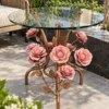 Round transparent glass side table with pink metal roses and bronze-gold floral legs in a sunlit seating area