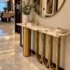 Luxury marble console table with gold cylindrical and U-shaped loop supports beneath a round mirror in a modern entryway.