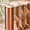 Close-up of white marble console tabletop with black and copper-toned veining beside cylindrical copper stainless steel and U-shaped loop supports.