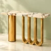 Gold stainless steel console table with rectangular white marble top, cylindrical pedestal and three U-shaped legs against a pale green background.