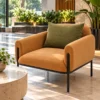 Modern caramel lounge chair with rounded upholstered armrests and olive-green cushion beside large windows and indoor plants.