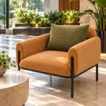 Modern caramel lounge chair with rounded upholstered armrests and olive-green cushion beside large windows and indoor plants.
