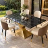 Black marble dining table with sculptural gold stainless steel base and cream dining chairs beside a garden-facing glass doorway.