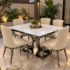 Six seater square white marble dining table with curved stainless steel pedestal and cream upholstered chairs in a garden-side patio setting.